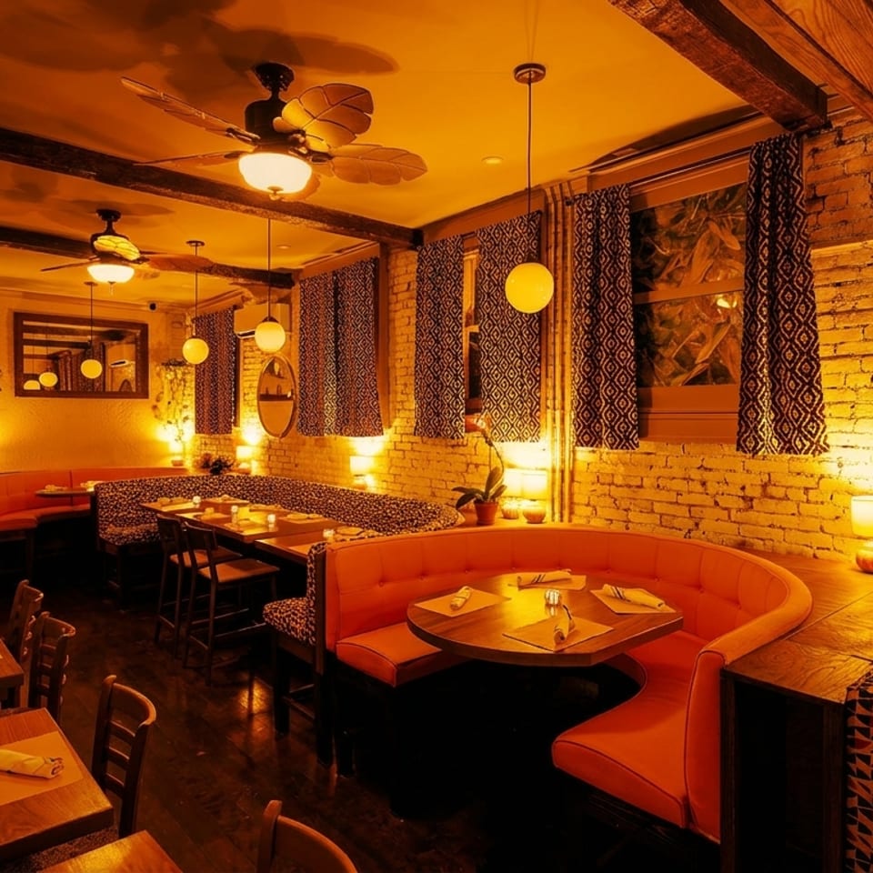 Warmly lit restaurant interior with orange booths, wooden tables, exposed brick, and patterned curtains.