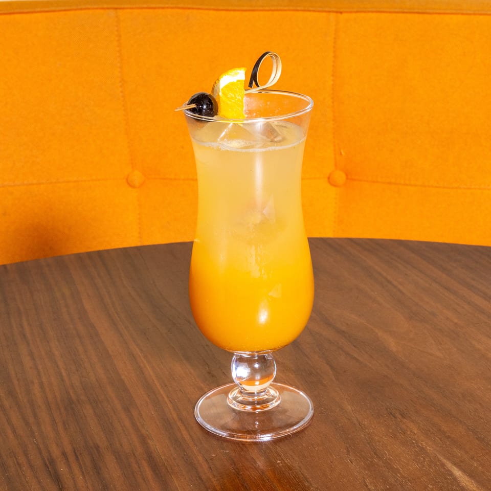 Orange cocktail with ice, orange slice, and cherry garnish served in a stemmed glass on a wooden table.