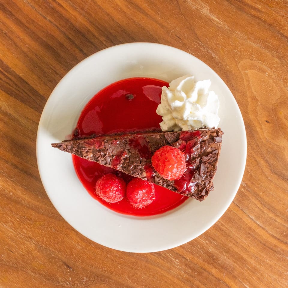 Chocolate cake slice topped with raspberries and whipped cream, served over raspberry sauce on a white plate.