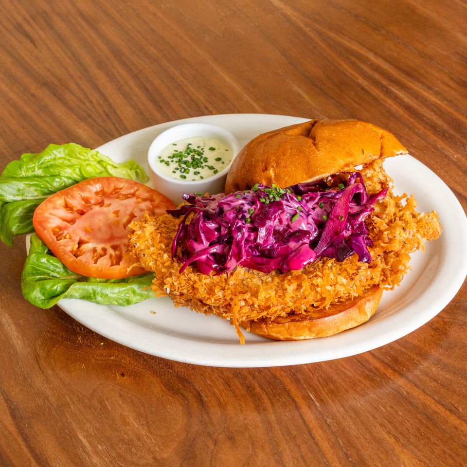 Crispy fried chicken sandwich topped with purple cabbage slaw on a plate with lettuce, tomato, and dipping sauce.