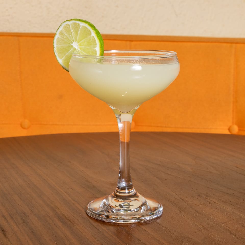 Creamy pale cocktail in a coupe glass garnished with a lime wheel on a wooden restaurant table.