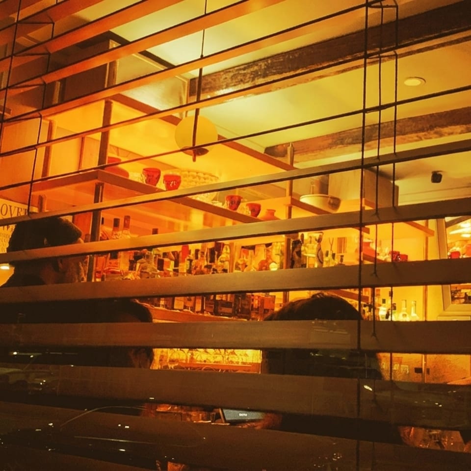 Warmly lit bar interior with bottles and patrons, viewed through horizontal blinds at night.