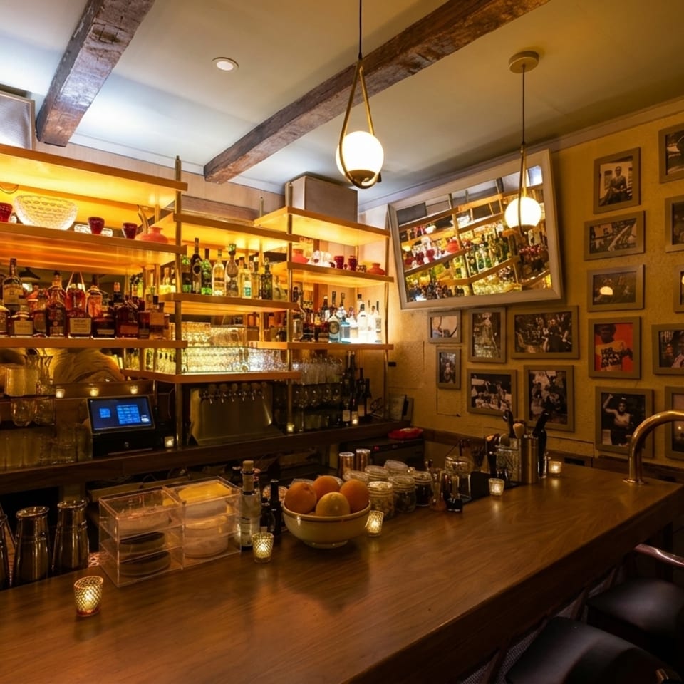 Warmly lit bar with shelves of liquor bottles, a wooden counter, and framed pictures on the wall.