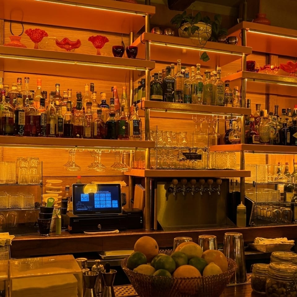 Warmly lit restaurant bar stocked with liquor bottles, glassware, cocktail ingredients, and colorful decorative shelves.