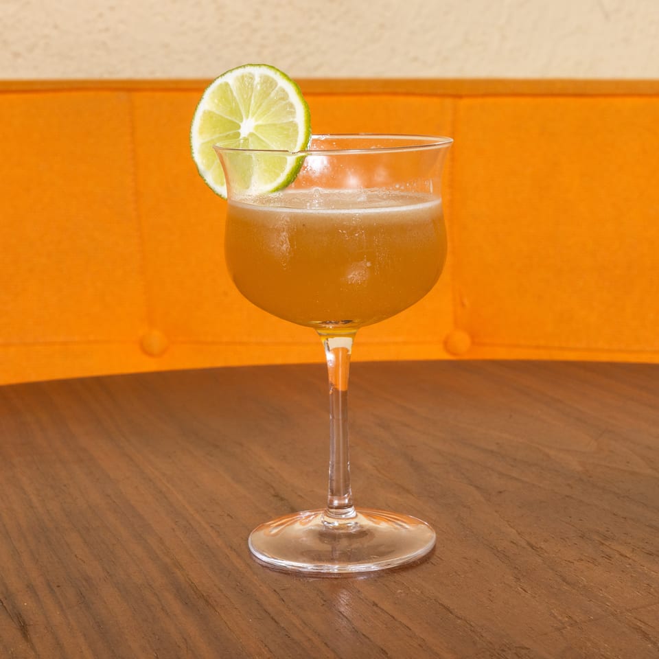 Golden cocktail in a stemmed glass garnished with a lime wheel, resting on a wooden table against an orange banquette.