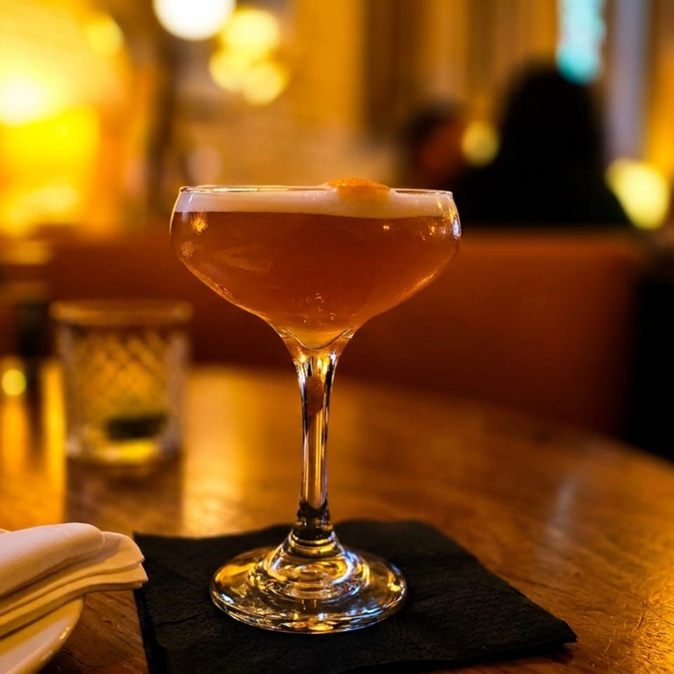 A coupe glass with an amber cocktail and orange peel sits on a dark table in a dimly lit restaurant.