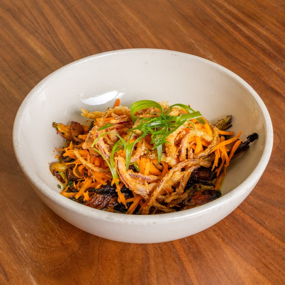 White bowl of crispy fried vegetables, shredded carrots, and green onion garnish on a wooden table.