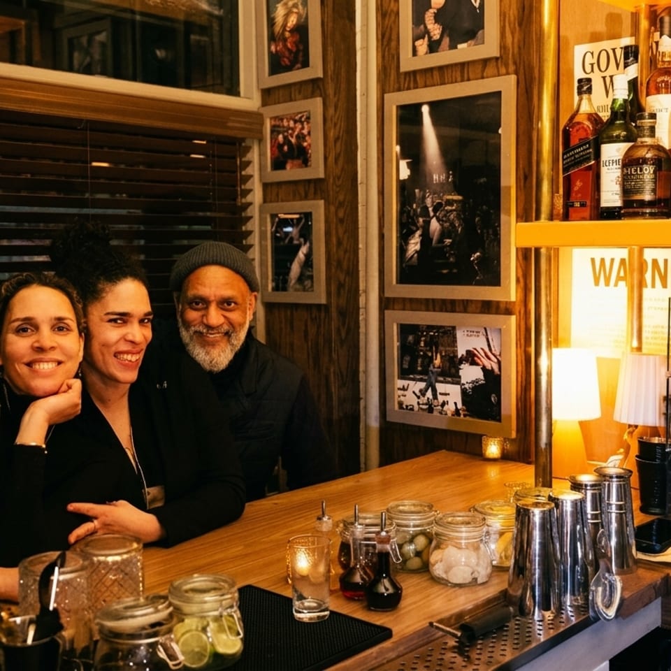 Three smiling people, two women and a man, pose behind a well-stocked bar.