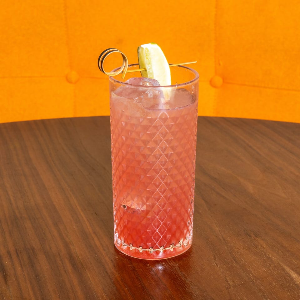 Pink cocktail with ice, a cucumber wedge, and decorative pick in a textured glass on a wooden table.
