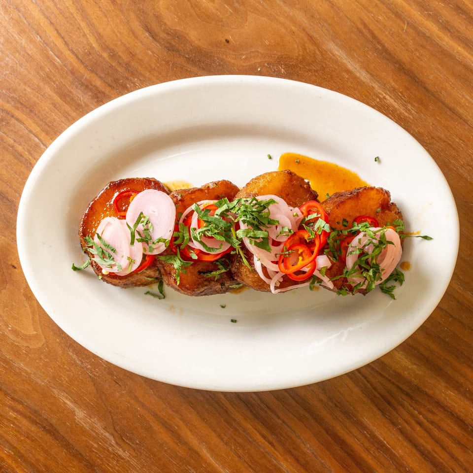 Roasted meat topped with pickled onions, red chilies, and fresh herbs on a white plate.