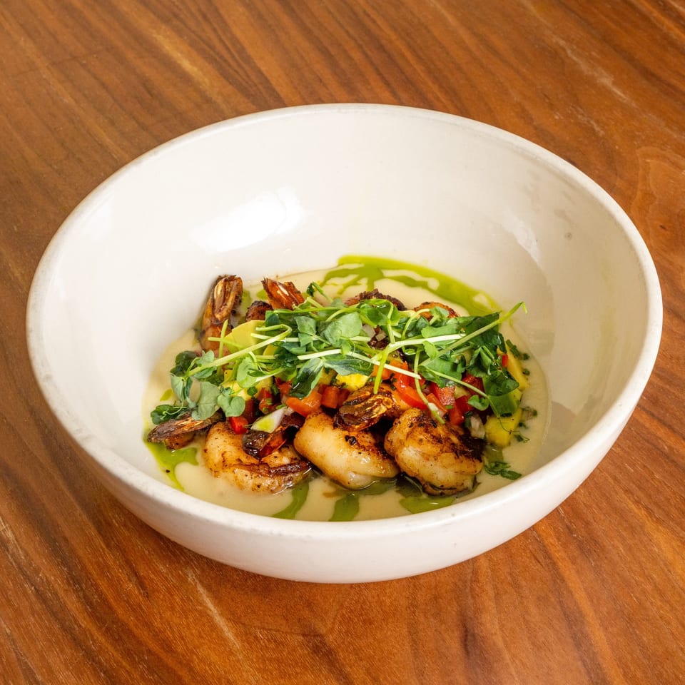 Seared scallops and shrimp topped with microgreens, diced vegetables, and green herb oil in a white bowl.