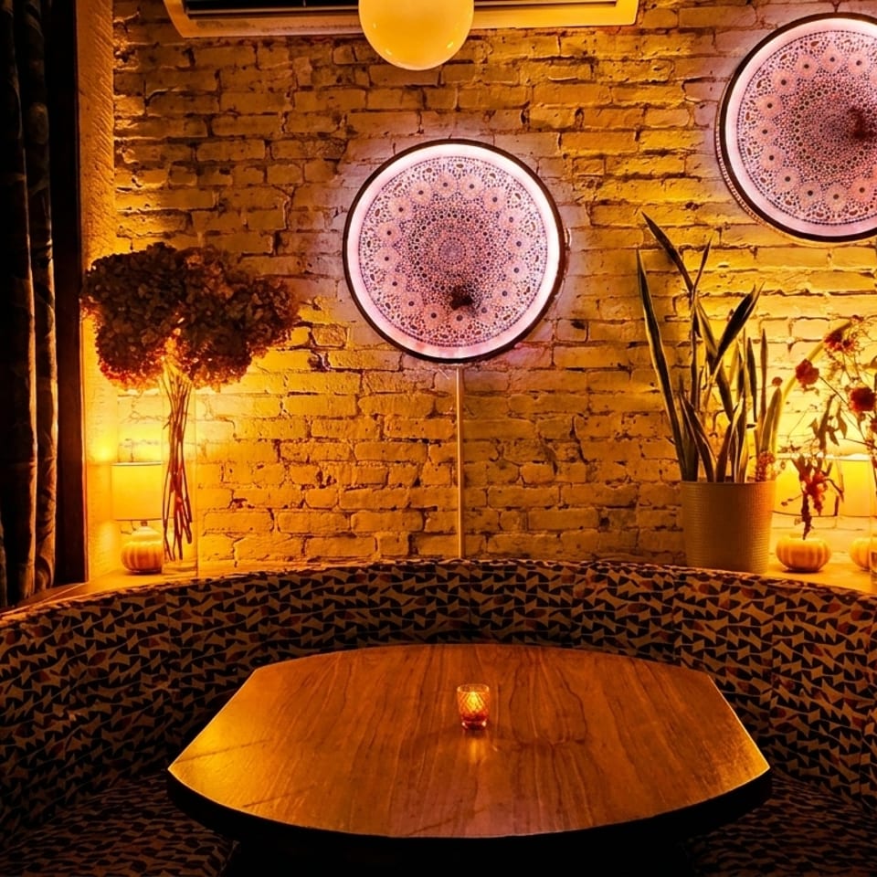 Cozy restaurant interior with patterned booth seating, wooden table, brick wall, and decorative lighting.
