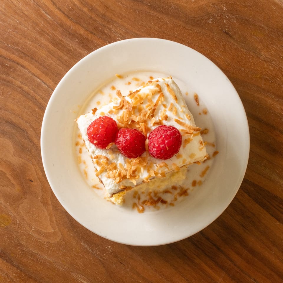 Slice of cream-topped cake garnished with raspberries and toasted coconut on a white plate.